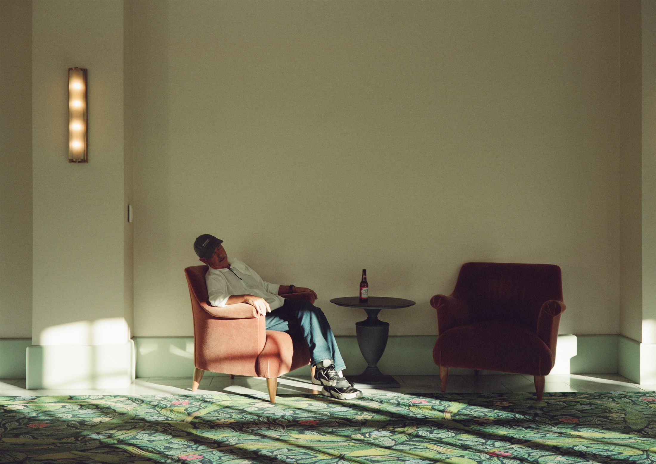 A man in a baseball cap resting in an armchair beside a floral carpet, with a bottle of beer on a side table.
